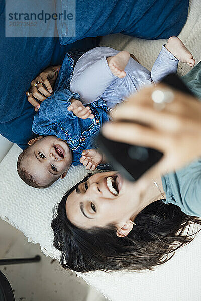 Cheerful mother taking selfie on smart phone with baby girl on bed
