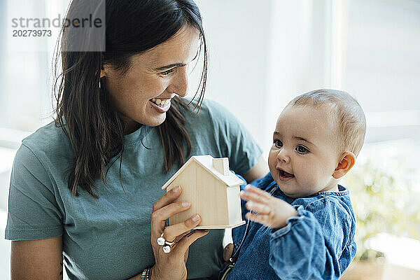 Happy mother showing wooden model home to baby daughter