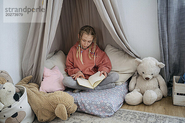 Mädchen mit Zöpfen liest zu Hause ein Buch im grauen Baldachin