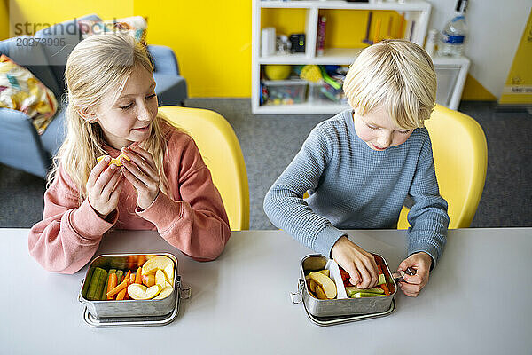 Blonder Junge und Mädchen essen Obst und Gemüse in der Lunchbox am Schreibtisch