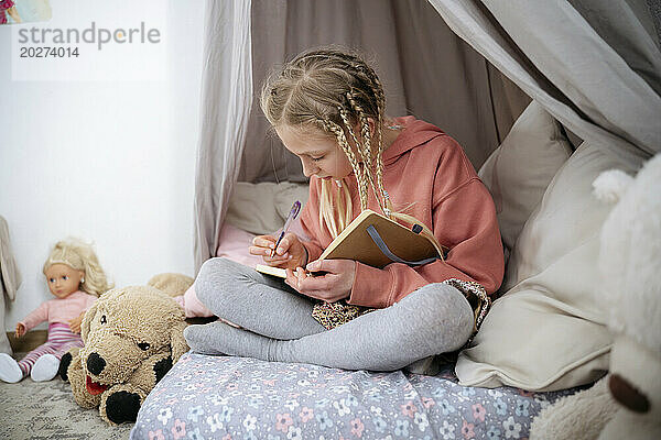Mädchen mit Zöpfen schreibt zu Hause im grauen Baldachin in ein Buch