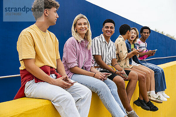 Fröhliche Freunde in farbenfroher Kleidung sitzen an einer gelben Wand und halten Smartphones in der Hand
