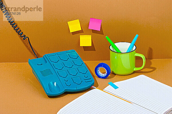 Blaues Telefon in der Nähe eines offenen Buches mit Büromaterial vor orangefarbenem Hintergrund