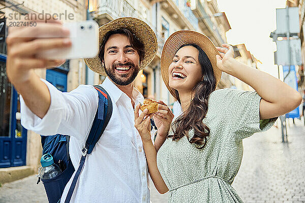 Lächelndes junges Paar mit Hut und Selfie mit traditionellem Dessert Pastel de Nata über das Smartphone