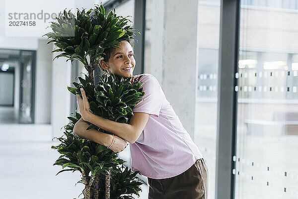 Lächelnde junge Geschäftsfrau umarmt Pflanze im Büro