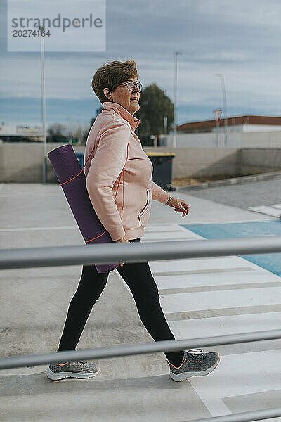 Aktive ältere Frau mit Gymnastikmatte geht auf Fußweg