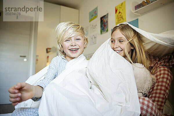 Happy brother and sister playing with blanket on bed at home