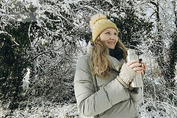 Lächelnde Frau hält Flasche in der Nähe von schneebedeckten Bäumen