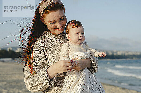 Smiling mother holding daughter at beach on sunny day