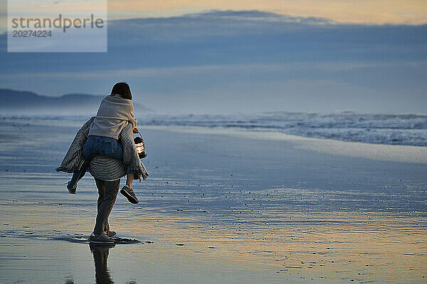Freund trägt Freundin huckepack und geht bei Sonnenuntergang am Strand spazieren