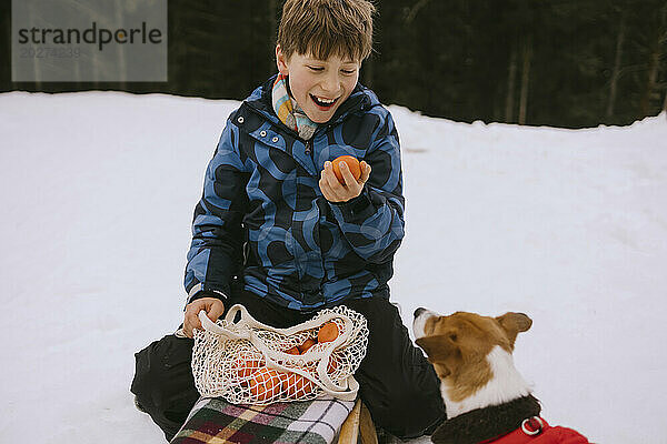 Glücklicher Junge hält Orange neben Hund im Winterwald