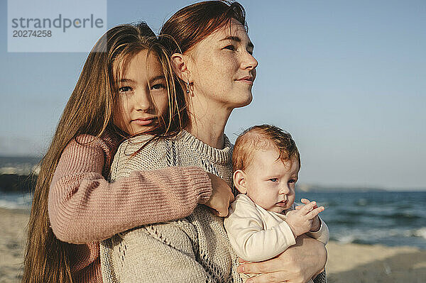 Frau hält an einem sonnigen Tag ein kleines Mädchen in der Nähe ihrer Tochter am Strand