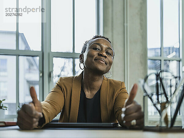 Selbstbewusste Geschäftsfrau zeigt im Büro die Geste „Daumen hoch“.