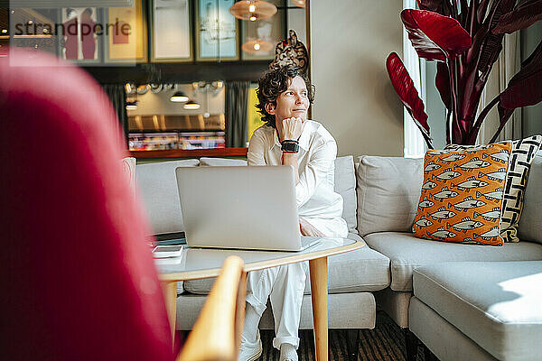 Nachdenkliche Geschäftsfrau sitzt mit der Hand am Kinn am Laptop in der Lobby des Hotels