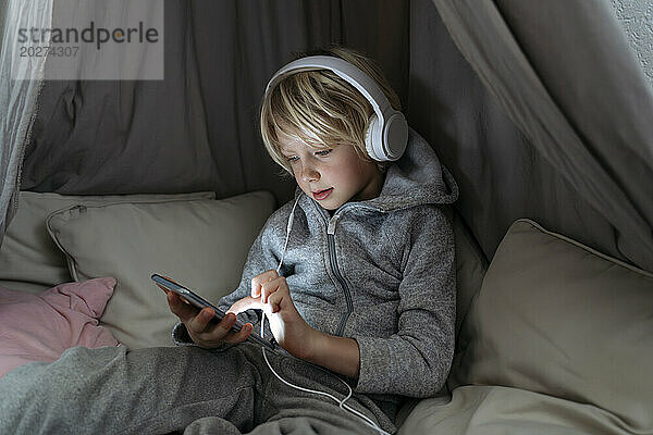 Junge mit Kopfhörern nutzt Internet auf Smartphone im Baldachin zu Hause