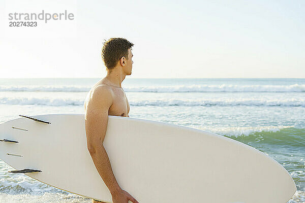 Junger Mann trägt Surfbrett und blickt aufs Meer