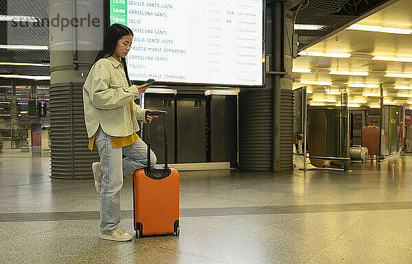 Junge Frau steht mit Koffer und benutzt Smartphone