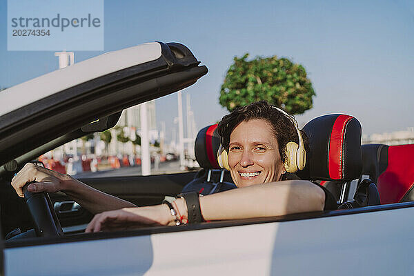 Lächelnde Frau mit kabellosen Kopfhörern im Cabrio an einem sonnigen Tag