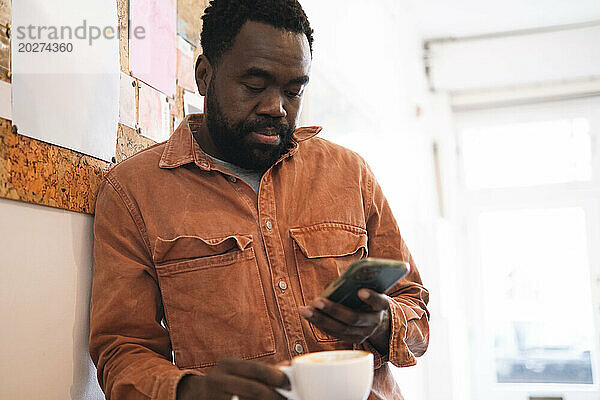 Mann hält Kaffeetasse in der Hand und benutzt Smartphone im Café