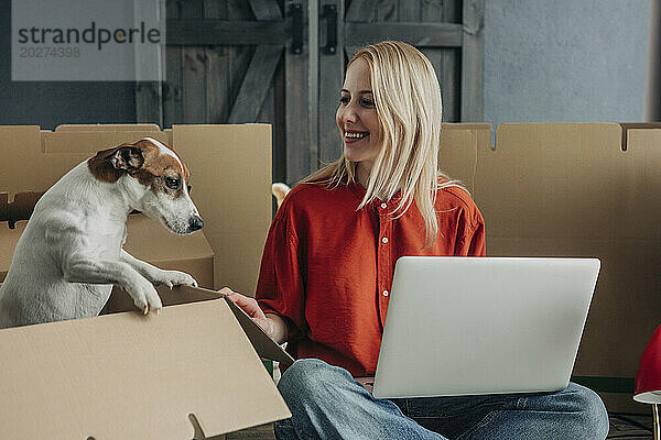 Lächelnde Frau sitzt mit Laptop neben Hund im Karton