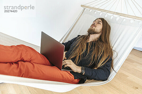 Junger Programmierer entspannt sich beim Start im Büro auf der Hängematte mit Laptop