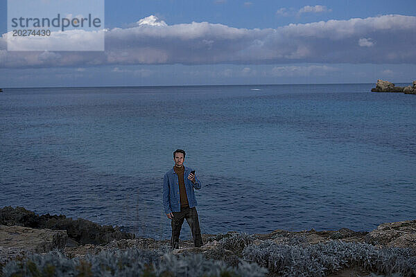 Mann mit Smartphone steht in der Abenddämmerung auf einem Hügel am Meer