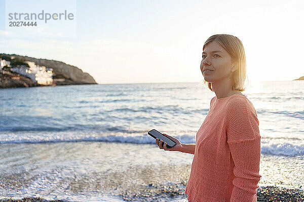 Nachdenkliche Frau mit Smartphone am Strand