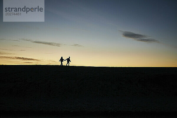 Silhouette eines jungen Paares  das Händchen hält und am Strand spaziert