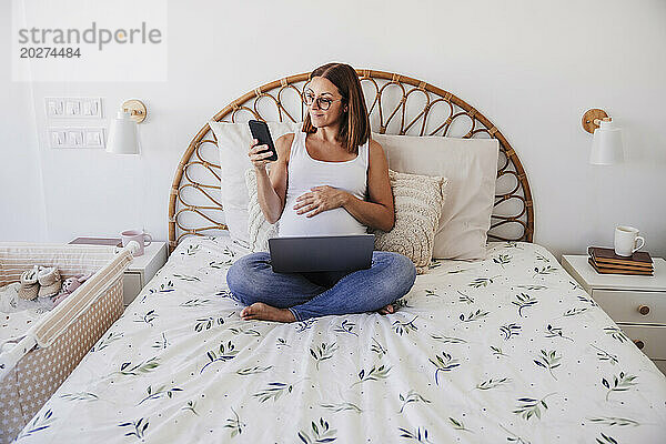 Schwangere Freiberuflerin nutzt Smartphone im Heimbüro im Bett