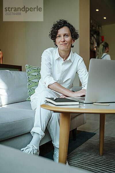 Lächelnde  selbstbewusste Geschäftsfrau mit Laptop sitzt auf dem Sofa in der Hotellobby