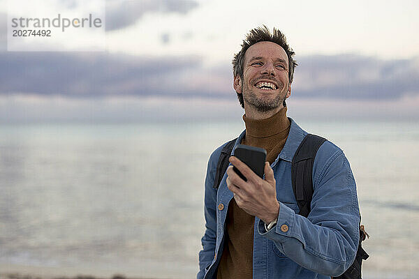 Sorgloser Mann mit Smartphone am Strand bei Sonnenuntergang