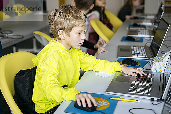 Blonder Junge trägt gelbes Sweatshirt und benutzt Laptop am Schreibtisch im Klassenzimmer