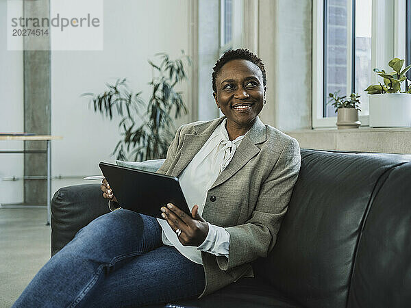 Lächelnde Geschäftsfrau im Blazer sitzt auf Sofa mit Tablet-PC im Büro