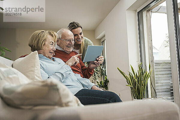 Glückliches Seniorenpaar und häusliche Pflegekraft nutzen Tablet-PC im Wohnzimmer