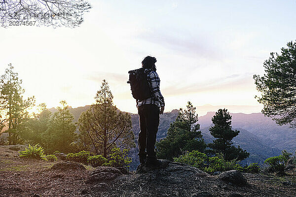Mann mit Rucksack steht bei Sonnenuntergang auf einem Felsen in der Nähe von Bäumen