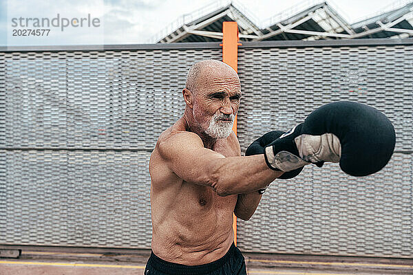 Oberkörperloser älterer Mann übt mit Boxhandschuhen am Zaun