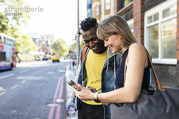 Lächelnde Frau teilt Smartphone mit Freund  der auf der Straße steht