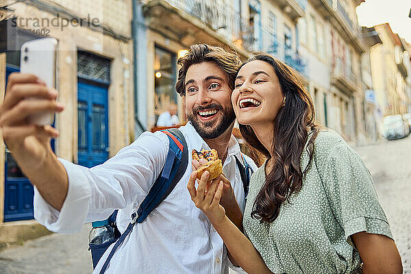 Fröhliches Paar hält traditionelles Dessert Pastel de Nata in der Hand und macht ein Selfie mit dem Smartphone