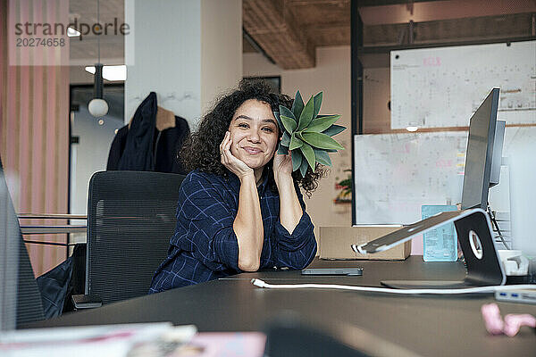 Lächelnde Geschäftsfrau hält Pflanze am Schreibtisch im Büro in Gesichtsnähe