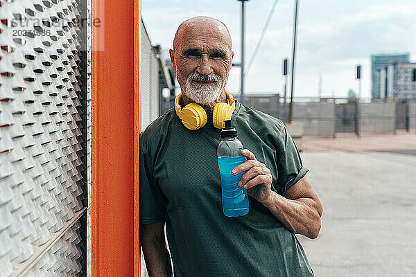 Aktiver älterer Mann mit kabellosen Kopfhörern hält Energy-Drink in der Hand und lehnt am Zaun