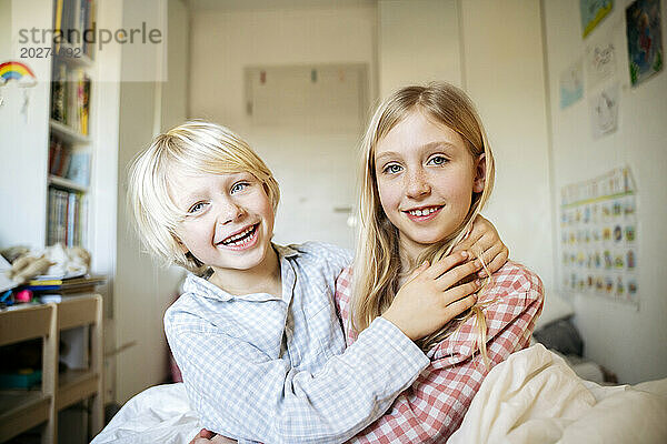 Smiling boy sitting with arm around sister in bedroom at home