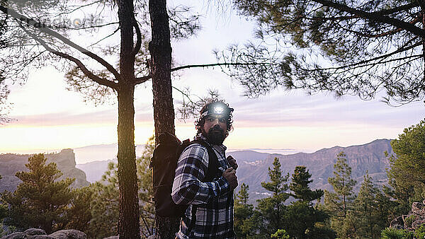 Mann trägt Stirnlampe und steht neben Bäumen vor Bergen