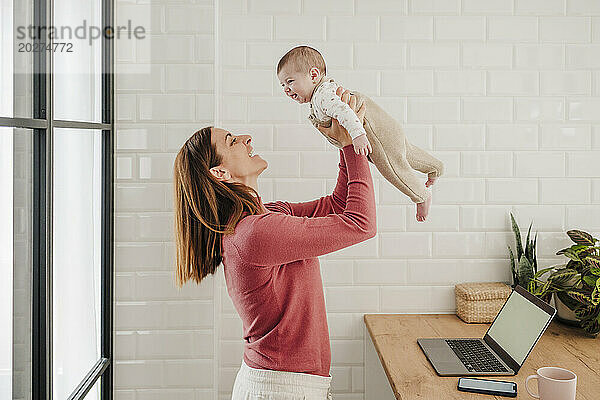 Happy freelancer mother playing with baby girl in kitchen at home