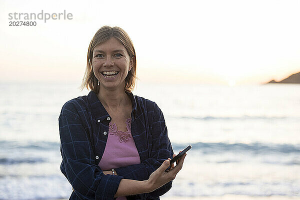 Fröhliche Frau mit Smartphone am Strand bei Sonnenuntergang