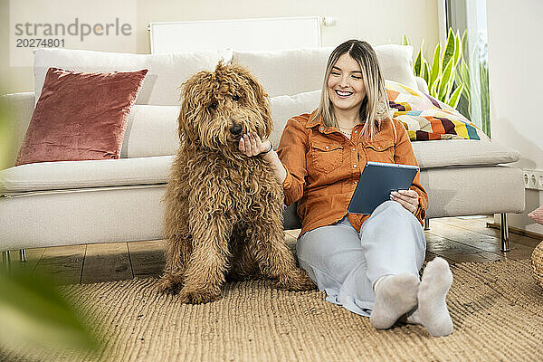 Glückliche schöne Frau hält Tablet-PC in der Hand und streichelt Hund zu Hause