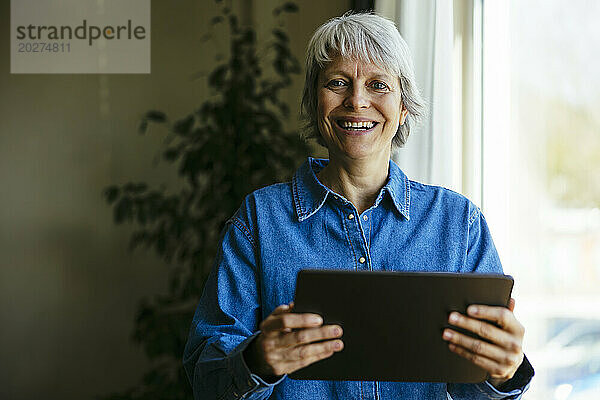 Glücklicher Freiberufler hält Tablet-PC am Fenster im Heimbüro