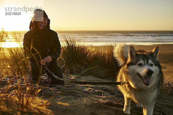 Fröhliche junge Frau spielt mit Husky-Hund am Strand