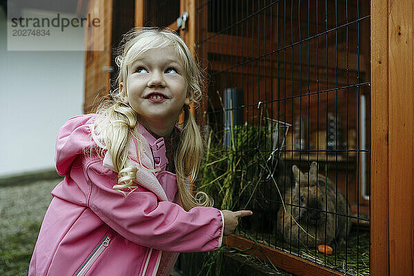 Blondes Mädchen im rosa Regenmantel füttert Kaninchen im Käfig