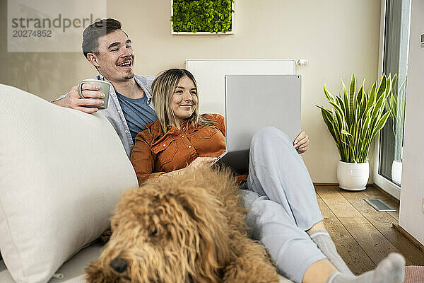 Glückliches Paar benutzt Laptop und sitzt zu Hause neben Hund