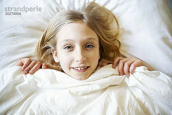 Smiling girl lying on bed with blanket at home